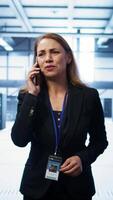 Vertical Software engineer talking on smartphone in data center, coordinating server maintenance tasks. Woman surrounded by server racks and high tech equipment, speaking during telephone call, camera B video