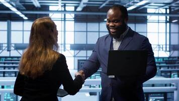African american admin greeting inspector arriving in data center to supervise operations. IT expert in server hub workspace doing hand shake with supervisor monitoring performance, camera A video