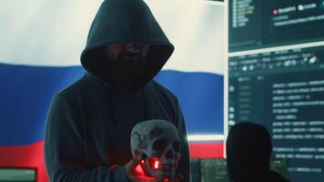 Conceptual image of a hooded russian hacker creating hazardous alerts, holds a skull and warning of death threats. Russian federation works on cybercrime and violence, panic and torment. Camera A. video
