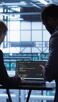 Vertical Team of developers doing maintenance checkup using laptop to prolong data center hardware life span. IT support staff members in server hub monitor server infrastructure using notebook, camera A video