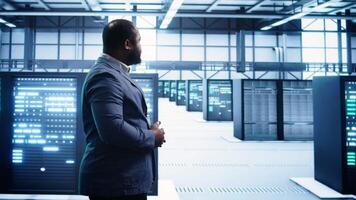 African american technician walking in data center, monitoring systems to identify errors. Man in server hub location doing diagnosis, looking for infrastructure bugs in need of repairing, camera B video