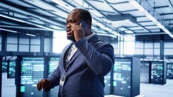 IT support worker in data center talking in phone call, doing yearly checkup. On field engineer chatting with manager in server hub during telephone call, monitoring servers infrastructure, camera B video