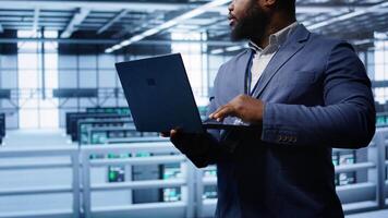 Computer scientist in server hub checking recovery plan on laptop, monitoring hardware energy consumption. IT specialist uses notebook in data center facility, ensuring rigs are functioning, camera B video