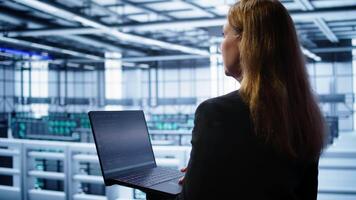System administrator in data center typing on laptop, running error checking utilities to fix problems. Woman in server hub looking for network issues affecting rigs using notebook device, camera B video