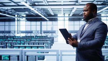 Developer in server hub checking recovery plan on tablet, monitoring hardware energy consumption. African american worker uses digital device in data center, ensuring rigs are functioning, camera B video