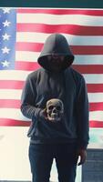 Vertical American man in a dark hood making death threats with human skull concept, emphasizing violence and malicious intent. Governmental hacker inflicting terror and panic for rival ideologies. Camera B. video