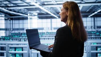 System administrator in server hub checking recovery plan on laptop, monitoring energy consumption. IT expert uses notebook in data center workspace, ensuring rigs are functioning optimally, camera B video