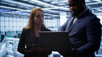 Team of IT specialists managing data center with advanced technology using devices. Coworkers in server room optimizing data center hardware equipment with laptop and tablet, camera B video