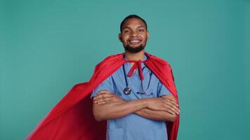 Smiling male nurse portraying superhero wearing red cape, isolated over studio background. Portrait of cheerful heroic medic posing as hero in costume with mantle, camera B video