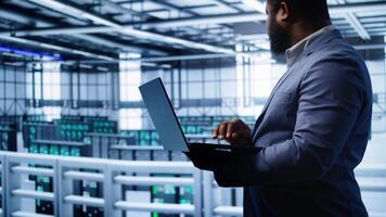 Software developer in server hub checking recovery plan on laptop, monitoring hardware energy consumption. African american worker uses notebook in data center, ensuring rigs are functioning, camera B video