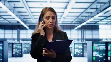 IT specialist looking over data center maintenance checklist, talking on phone in server farm. Employee surrounded by server racks talking during telephone call, camera B video