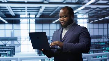 Programmer in data center typing on laptop, running error checking utilities. IT professional in server room looking for network issues affecting rigs performance using digital device, camera A video