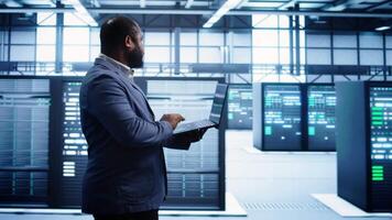 African american man walking in high tech facility, using laptop to patch network issues leading to slow data transfer rates. Data center worker fixing errors affecting supercomputers, camera B video