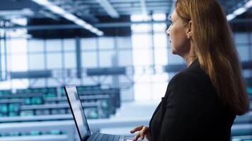 System administrator in data center typing on laptop, running error checking utilities to fix problems. Woman in server hub looking for network issues affecting rigs using notebook device, camera A video