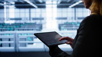 System administrator doing annual checkup using tablet to prolong data center equipment life span. Close up shot of IT expert in server hub monitoring supercomputers using digital device, camera B video
