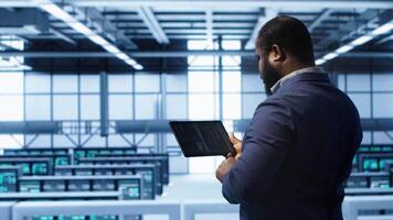 Computer scientist in data center running error checking utilities on tablet to fix bugs. IT expert in server hub facility looking for network issues affecting supercomputers performance, camera A video