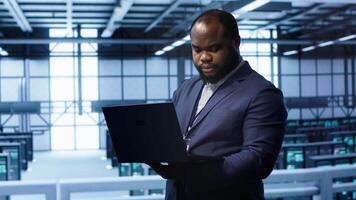 African american IT operator doing maintenance using laptop to prolong data center equipment life span. IT professional in server hub workspace monitoring server mainframes using notebook, camera A video