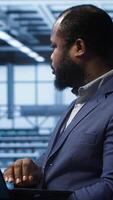 Vertical Software developer in server hub checking recovery plan on laptop, monitoring hardware energy consumption. African american worker uses notebook in data center, ensuring rigs are functioning, camera A video
