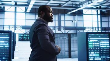 African american technician walking in data center, monitoring systems to identify errors. Man in server hub location doing diagnosis, looking for infrastructure bugs in need of repairing, camera A video