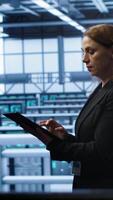 Vertical Computer scientist doing checkup, inputting data on tablet of needed fixes in server hub. Woman working in data center housing advanced data storage infrastructure supporting IT workloads, camera A video
