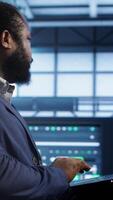 Vertical African american man walking in high tech facility, using laptop to patch network issues leading to slow data transfer rates. Data center worker fixing errors affecting supercomputers, camera A video