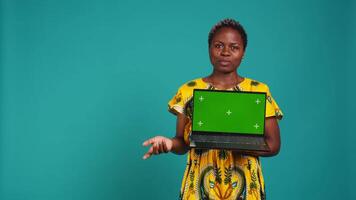 Young adult showing a laptop with isolated mockup screen in studio, presenting a display with green screen against blue background. Female model posing with chroma key. Camera B. video