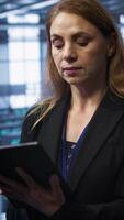 Vertical Admin in server room checks recovery plan on tablet, monitoring equipment energy consumption. IT professional uses notebook in data center workplace, ensuring rigs are functioning optimally, camera A video