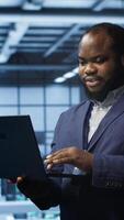Vertical Programmer in data center typing on laptop, running error checking utilities. IT professional in server room looking for network issues affecting rigs performance using digital device, camera A video