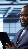 Vertical Programmer in server room location using tablet to patch network issues affecting transfer rates. African american man in data center fixing errors affecting mainframes, camera A video
