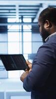 Vertical Computer scientist in data center running error checking utilities on tablet to fix bugs. IT expert in server hub facility looking for network issues affecting supercomputers performance, camera A video