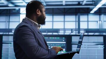 African american man walking in high tech facility, using laptop to patch network issues leading to slow data transfer rates. Data center worker fixing errors affecting supercomputers, camera A video