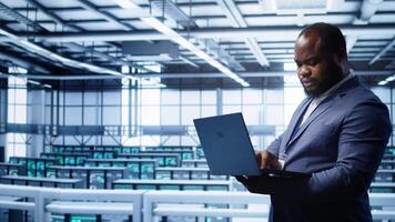 African american IT operator doing maintenance using laptop to prolong data center equipment life span. IT professional in server hub workspace monitoring server mainframes using notebook, camera B video