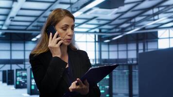 IT specialist looking over data center maintenance checklist, talking on phone in server farm. Employee surrounded by server racks talking during telephone call, camera A video