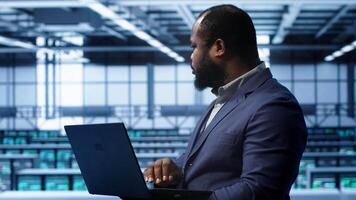 Software developer in server hub checking recovery plan on laptop, monitoring hardware energy consumption. African american worker uses notebook in data center, ensuring rigs are functioning, camera A video