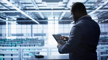 Technician in server hub workplace installing newly ordered storage unit, checking parameters with tablet. IT expert in data center holding device, replacing storage mainframes components, camera B video