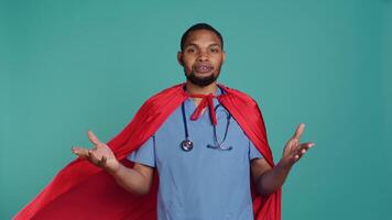 Male nurse portraying superhero showing courage and strength fighting diseases, studio backdrop. Portrait of african american doctor posing as hero in costume, holding empowering speech, camera B video