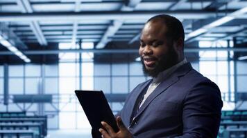 Programmer in server room location using tablet to patch network issues affecting transfer rates. African american man in data center fixing errors affecting mainframes, camera A video