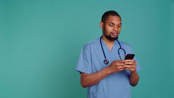 Male nurse using mobile phone to scroll on social media feed during hospital job shift break. Healthcare worker using smartphone to text friends, studio backdrop, camera B video