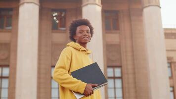 Young smiling black programmer in casual clothes with copybooks in his hands standing and looking at the camera with a building as a backdrop video