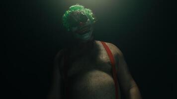Scary man in the horror clown mask with green hair, standing in suspenders in the dark studio with flashing lights, looking at the camera and making threatening gestures close up video