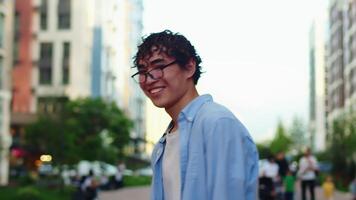 Young curly asian man in casual clothes and glasses walking on a city street, then turning around and smiling with multi-storey buildings as a backdrop video