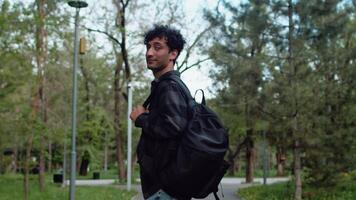 Young curly man in casual clothes with a backpack walking in an alley of the city park, then turning around and smiling. Concept of carefree urban life video
