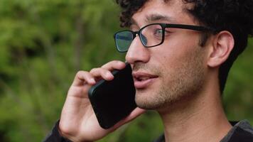 Young curly man in glasses standing in the city park, talking on a smartphone and cheerfully smiling close up. Concept of a relaxed conversation with a friend video