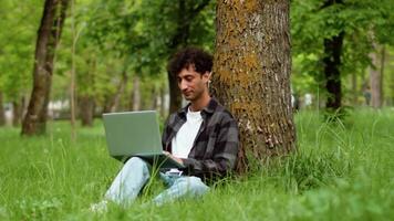 Young curly man in casual clothes sitting with a laptop under the tree in a city park and working outdoors with a smiling face. Concept of flexible remote work video