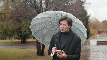 Young man with an annoyed face in a coat under an umbrella standing in the autumn city park in rainy weather, using his smartphone and looking around video