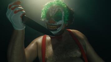 Scary man in the horror clown mask with green hair, standing in suspenders in the dark studio with flashing lights and threatening with the knife close up video