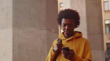 Young black programmer in casual clothes walking alongside the building outdoors, looking at the smartphone and smiling after a successful working day video