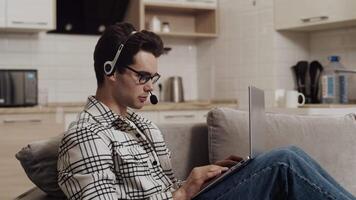 Young caucasian man in a headset sitting on a couch at home with a laptop on his knees, typing and talking with a client during a support service session video