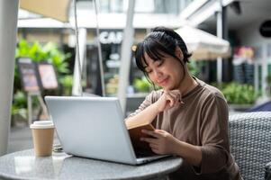 un mujer es trabajando remotamente a un cafetería, sentado exterior, utilizando su ordenador portátil y leyendo un libro. foto