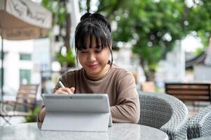un feliz, positivo asiático mujer es trabajando de forma remota, utilizando su digital tableta a un al aire libre salón. foto